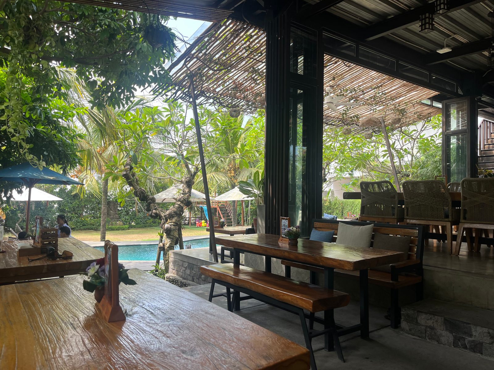 A shaded open-sided terrace with long wooden tables looking onto a pool and a tropical garden