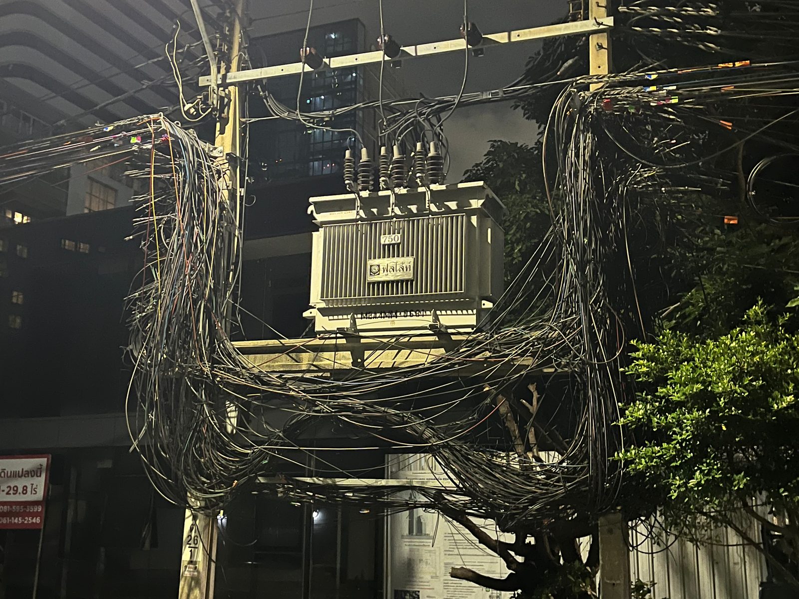 A thick tangle of overhead cables around a transformer on a Bangkok street at night