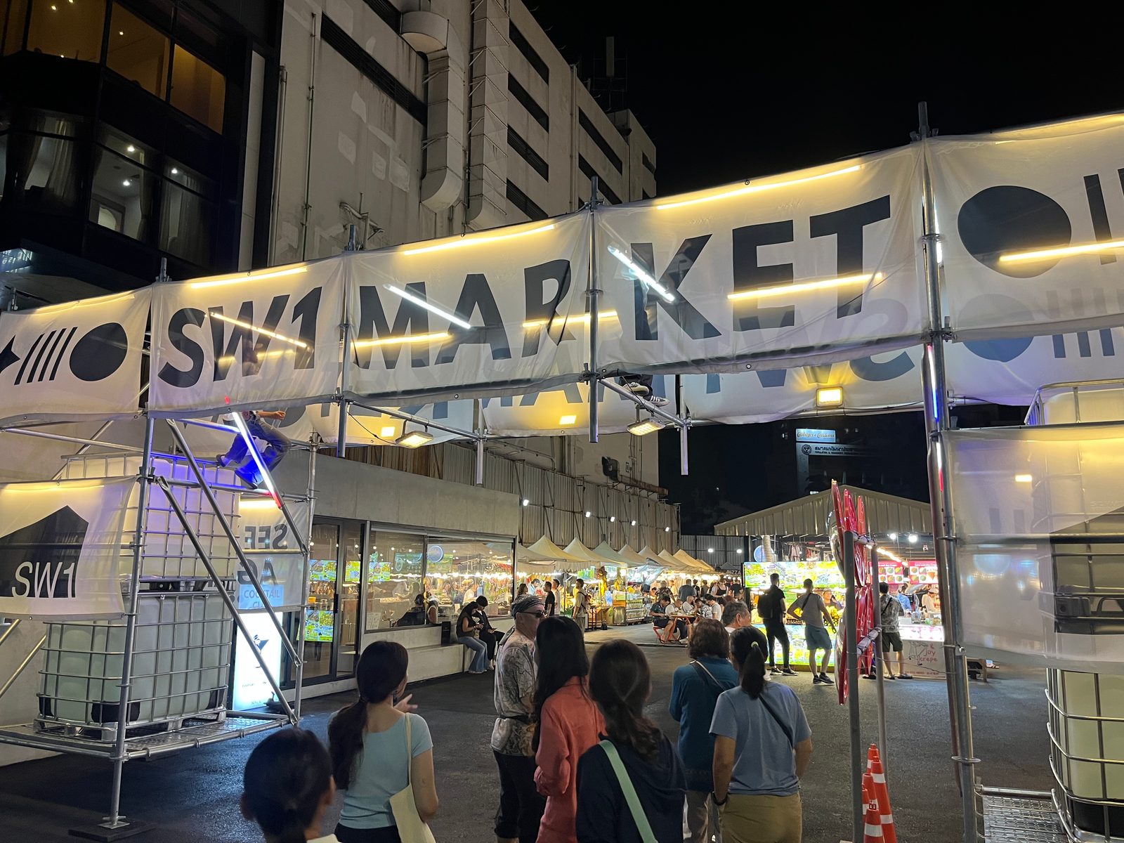 The lit entrance of SW1 Market at night, with people walking in toward the stalls