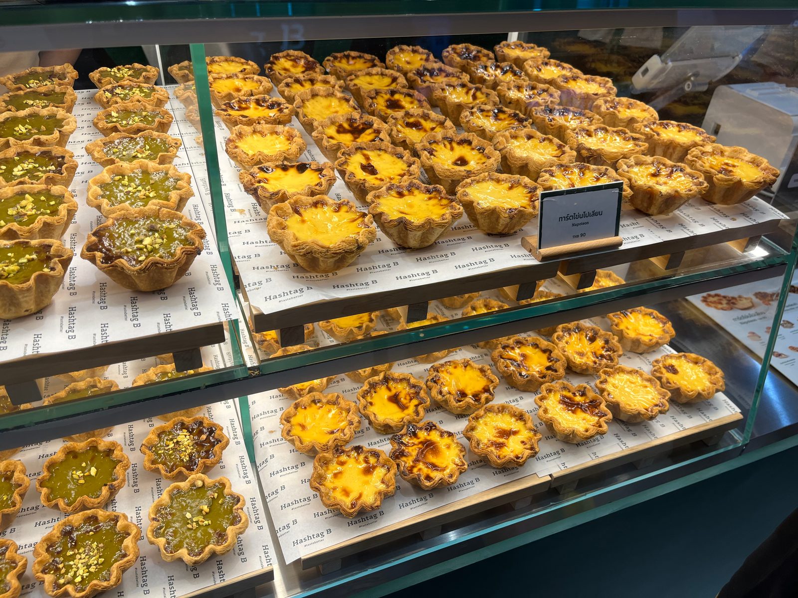 Rows of egg tarts and green custard tarts in a bakery display case at Siam Paragon
