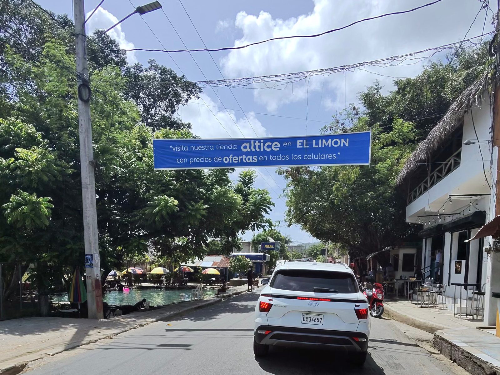 Blue Altice banner hung across a village street advertising its store in El Limón, with a white SUV ahead