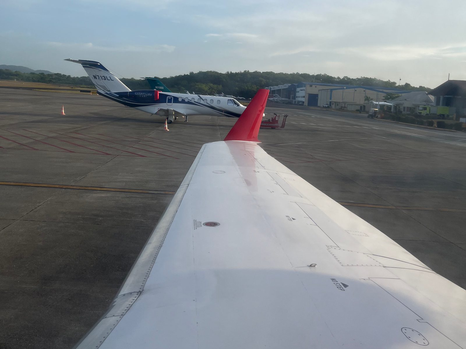 Aircraft wing with a red winglet seen from the cabin window, with a private jet parked on the apron at La Isabela airport