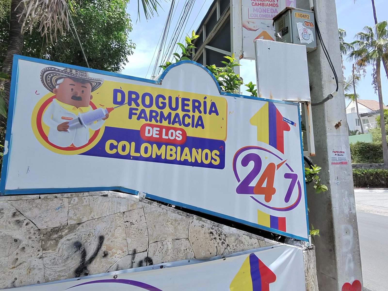 Roadside sign for the Droguería Farmacia de los Colombianos advertising 24/7 service, beside a utility pole and palm trees