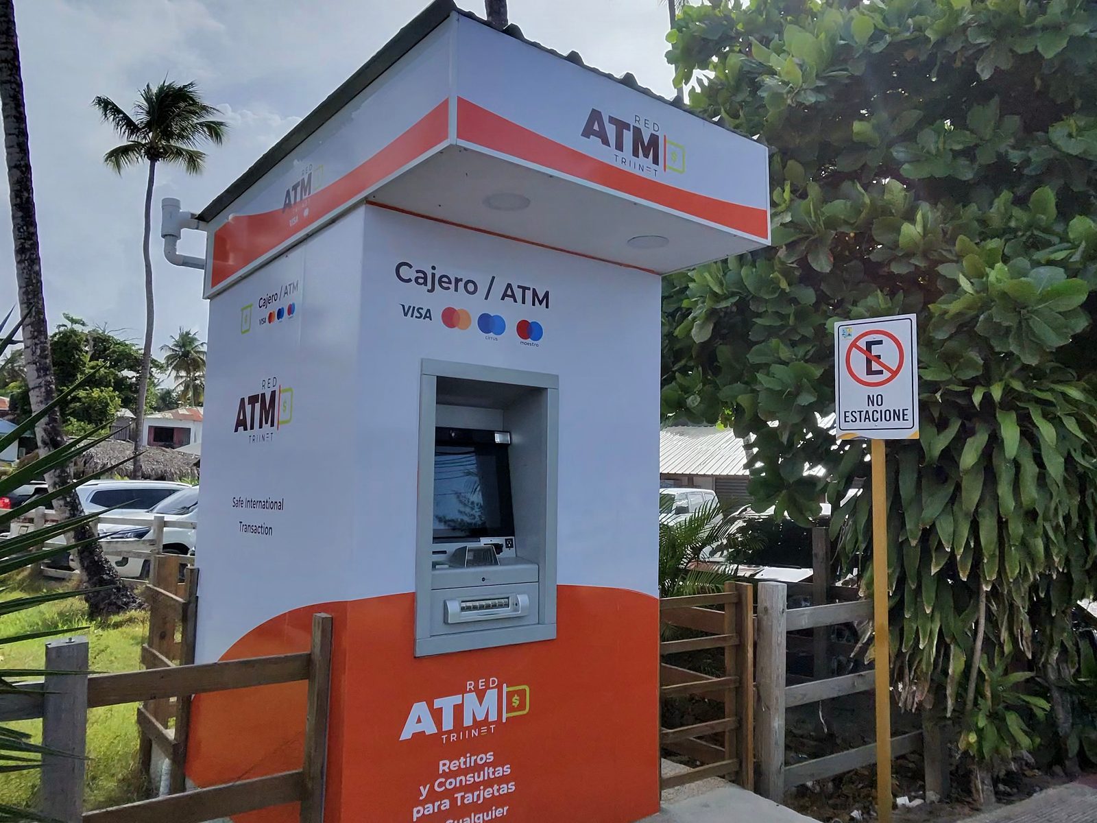 Standalone orange and white ATM kiosk beside a road with palm trees