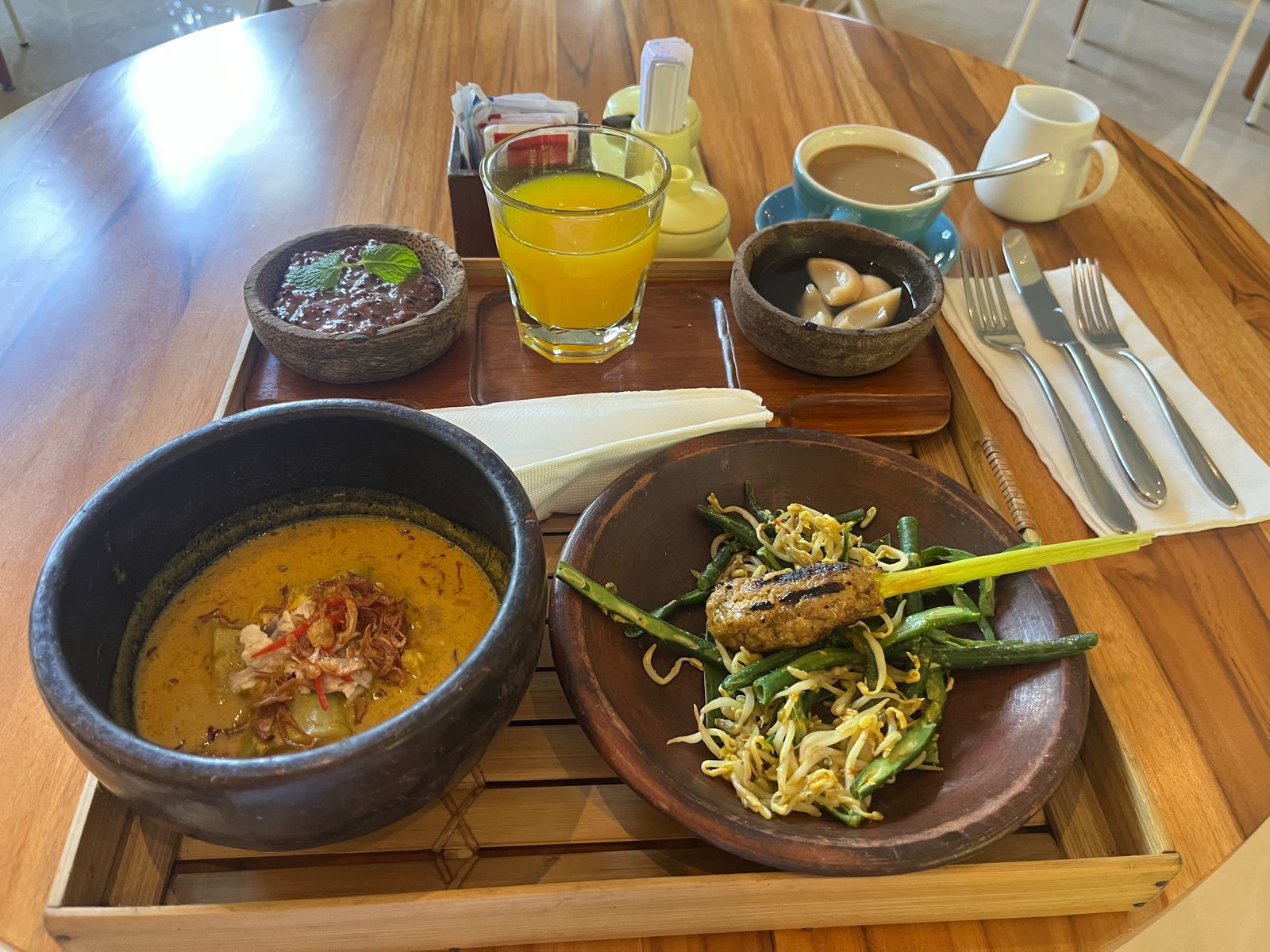 A breakfast tray with a bowl of yellow curry soup, green beans and bean sprouts with a lemongrass satay, small side bowls, orange juice and coffee