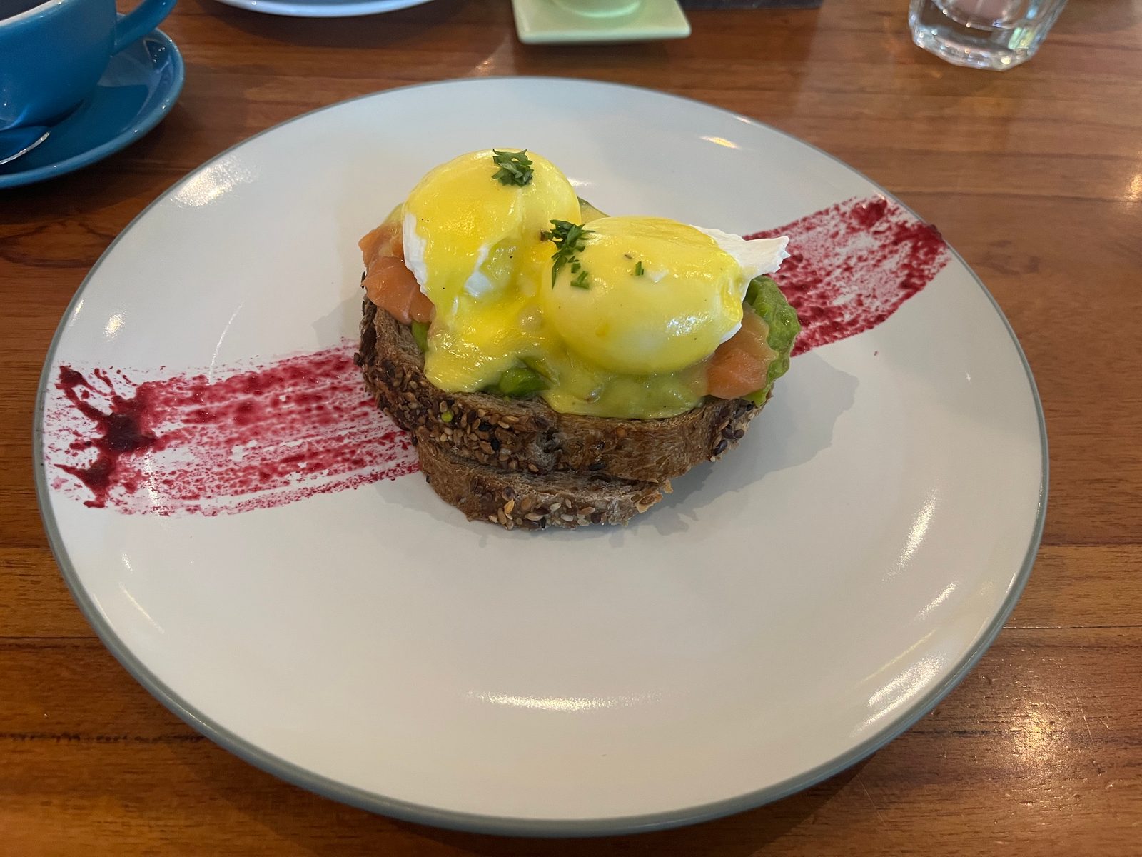Eggs Benedict with smoked salmon and avocado on seeded bread, with a streak of red sauce on the plate