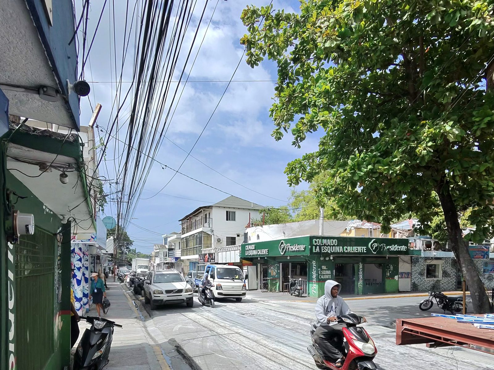 Dense bundle of overhead power and telecom cables above a street in central Las Terrenas, with a green colmado on the corner and a scooter passing