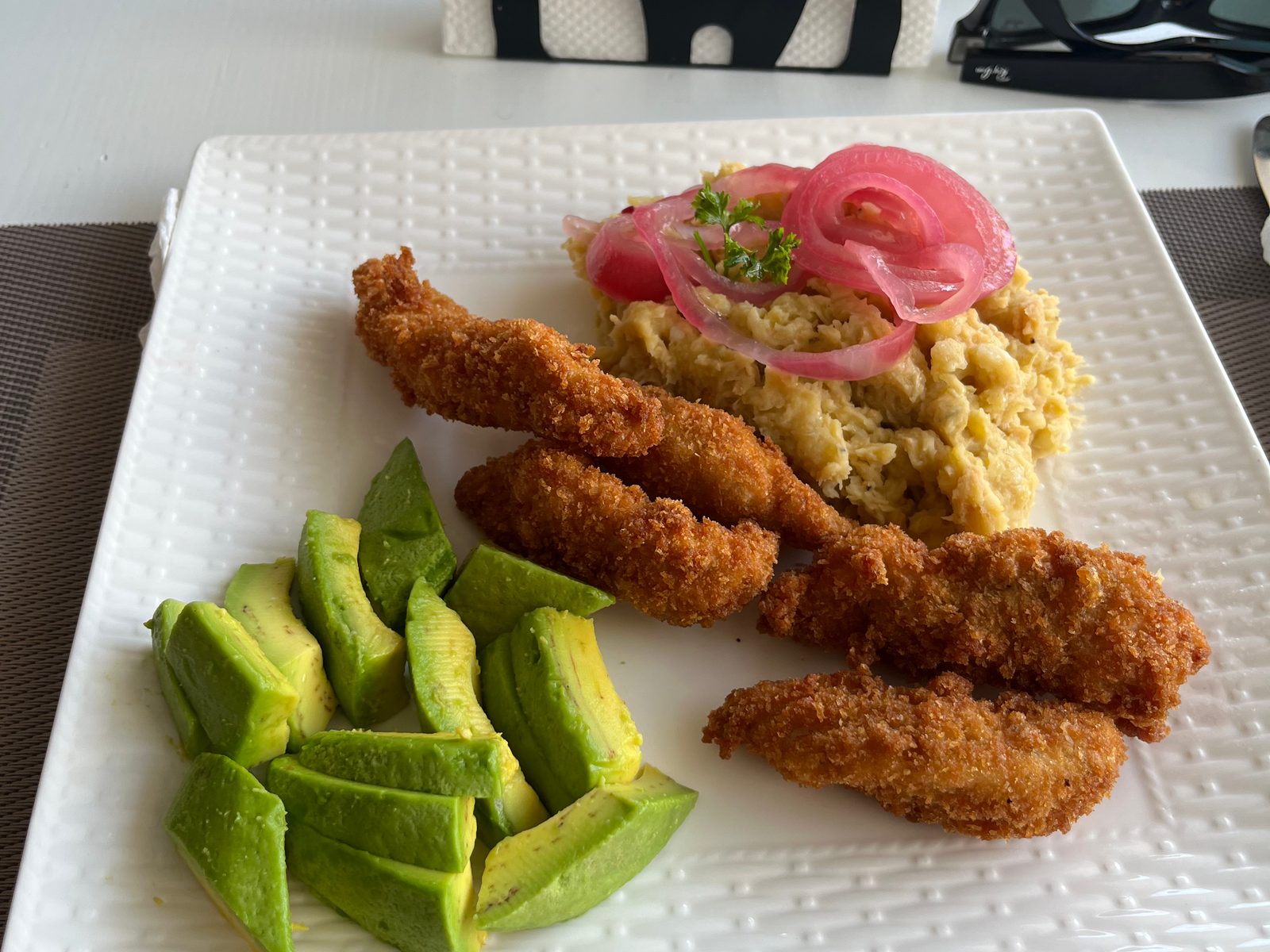 Mangú topped with pickled red onion, fried breaded strips and sliced avocado on a white plate
