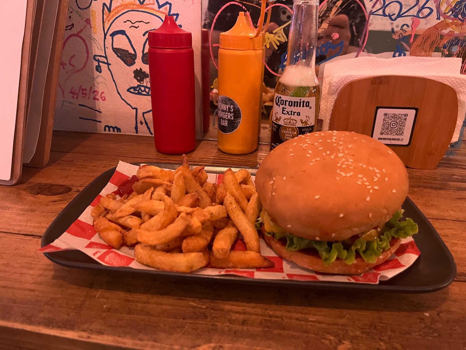 A burger and fries on a tray, with ketchup and mustard bottles, a Coronita and a QR code stand behind