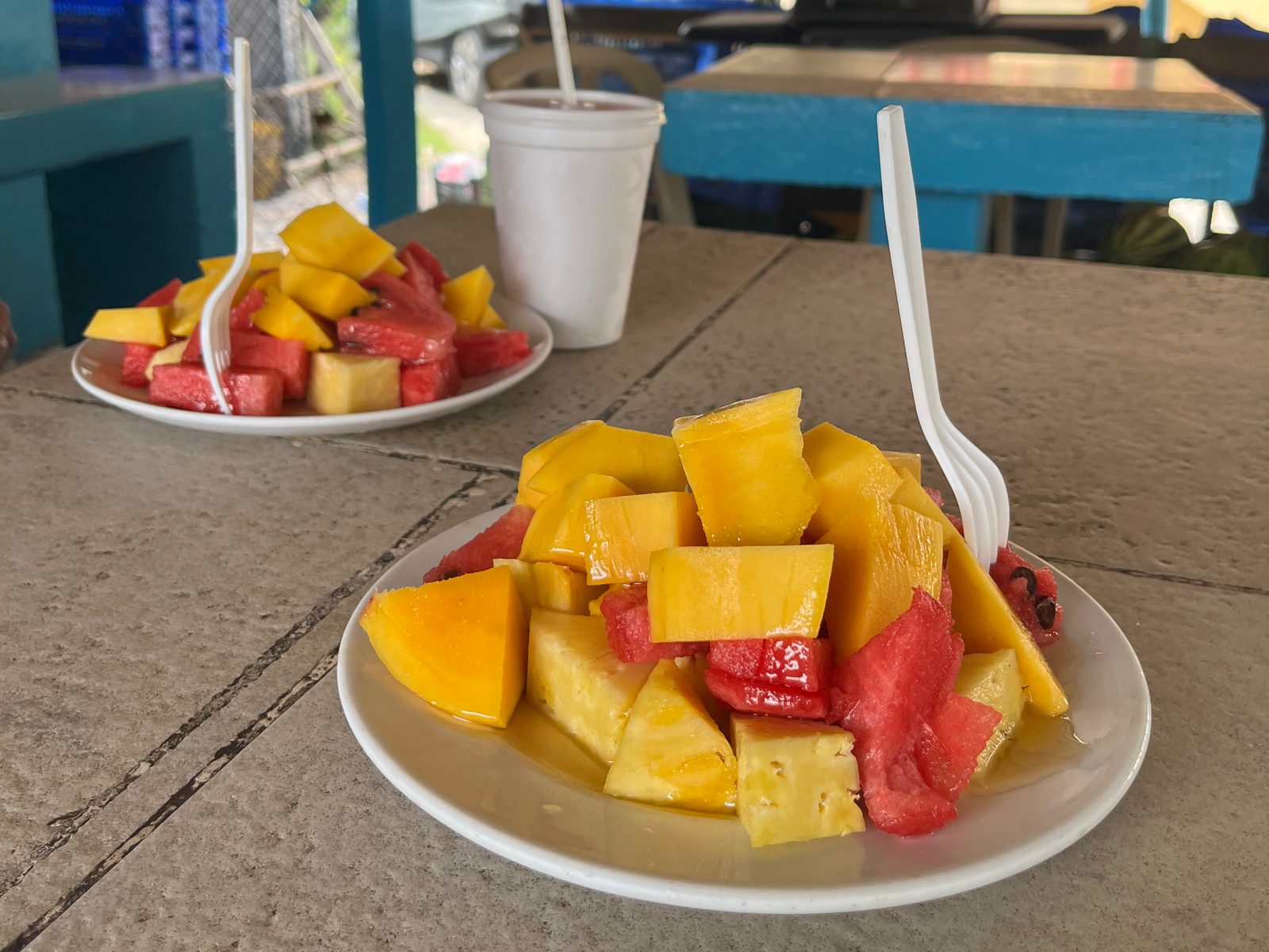 Two plates of cut mango, pineapple and watermelon on a table under a thatched roof
