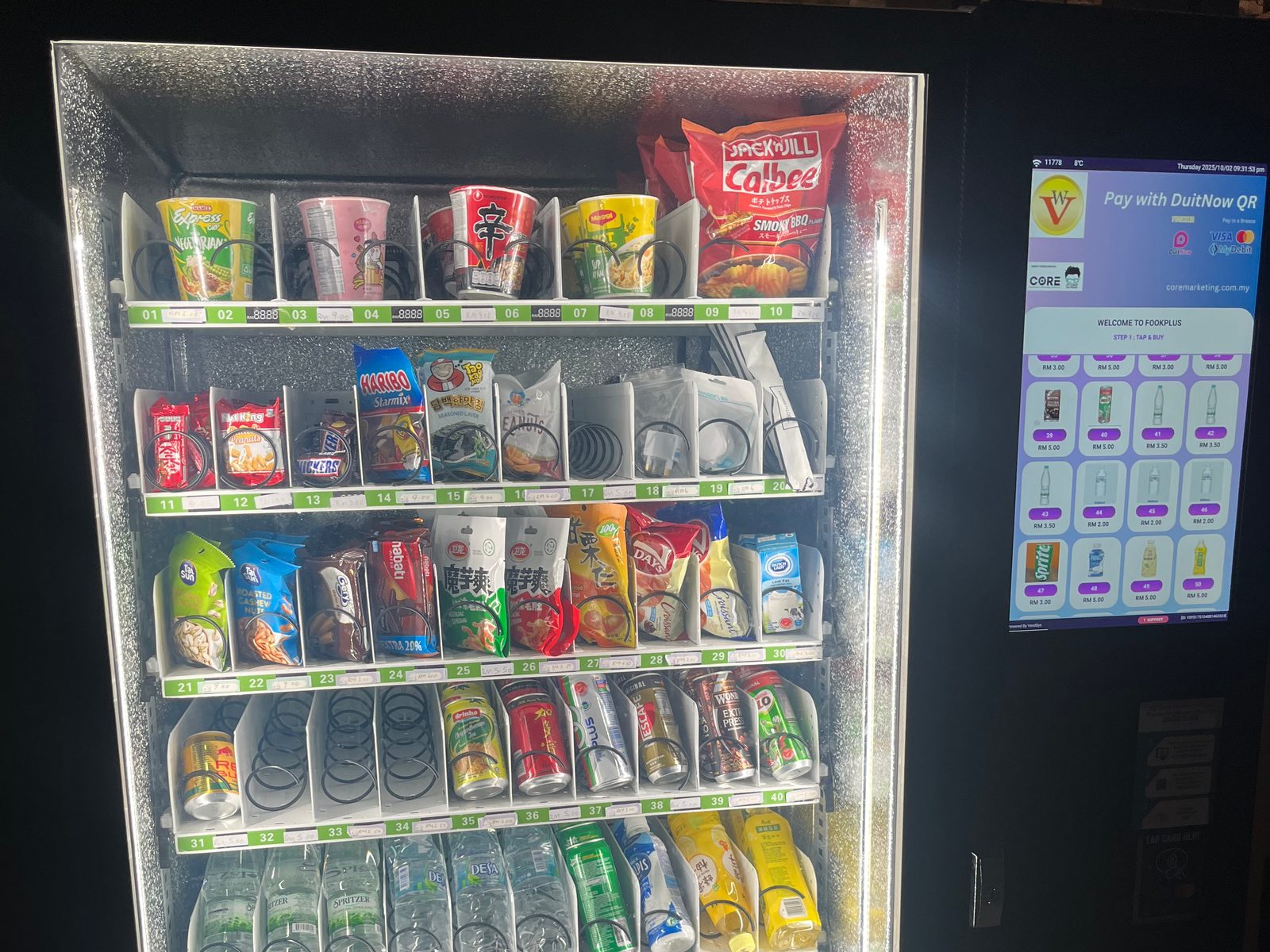 Lit vending machine with snacks and drinks next to a screen reading Pay with DuitNow QR