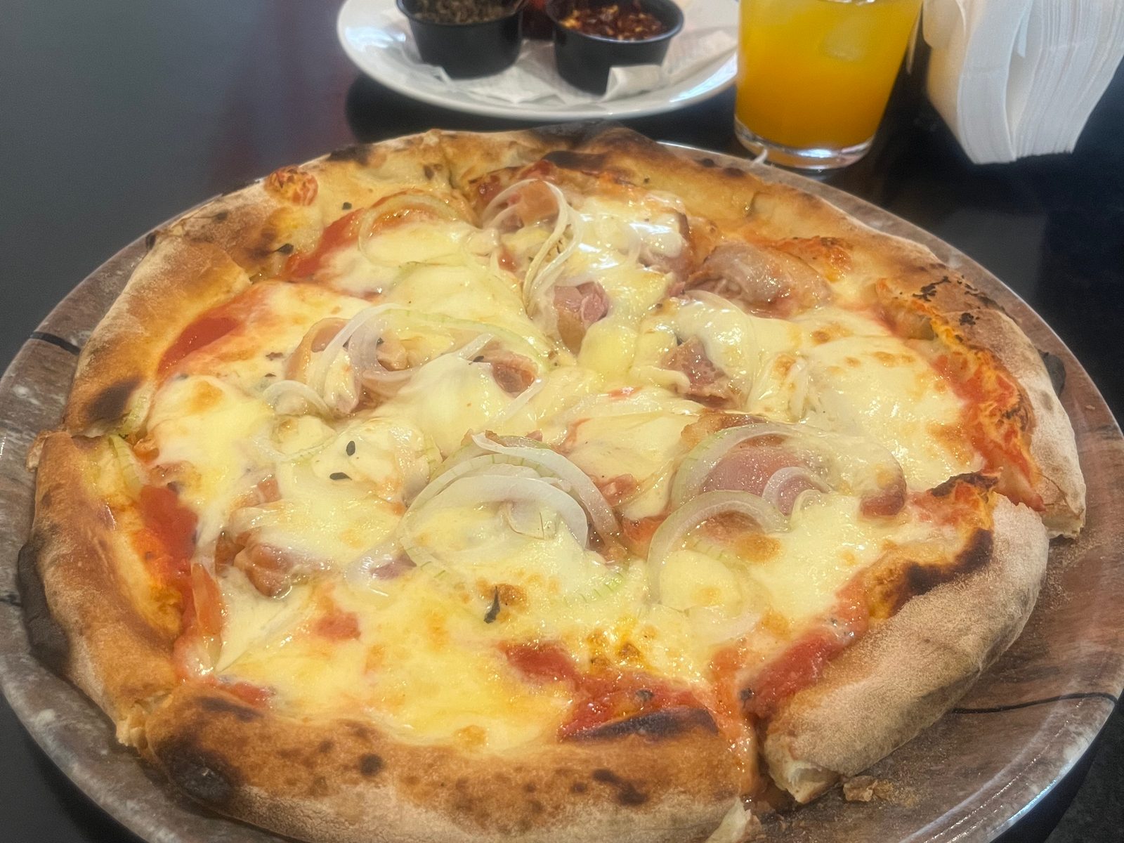 Pizza topped with cheese and onion on a metal tray, with a glass of juice and small bowls of hot sauce