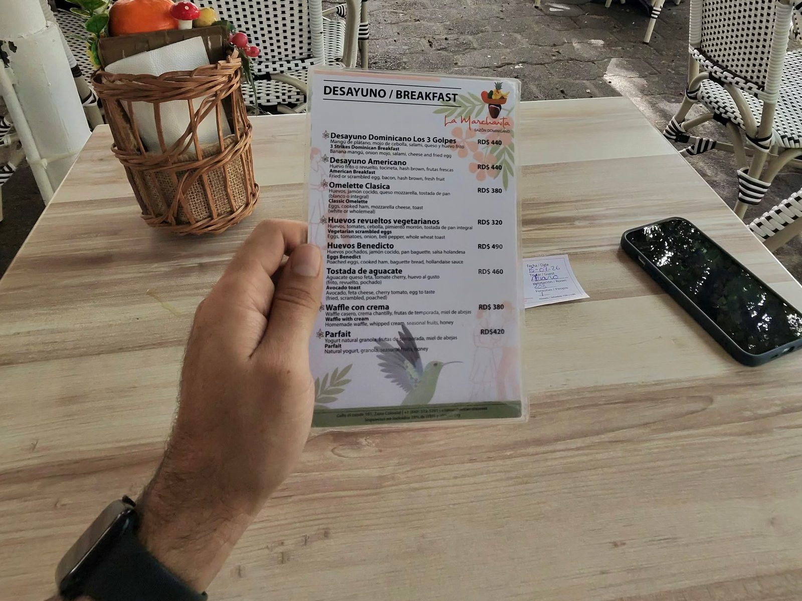 Hand holding a bilingual breakfast menu with prices in pesos over a café table, next to a phone and a wicker napkin holder