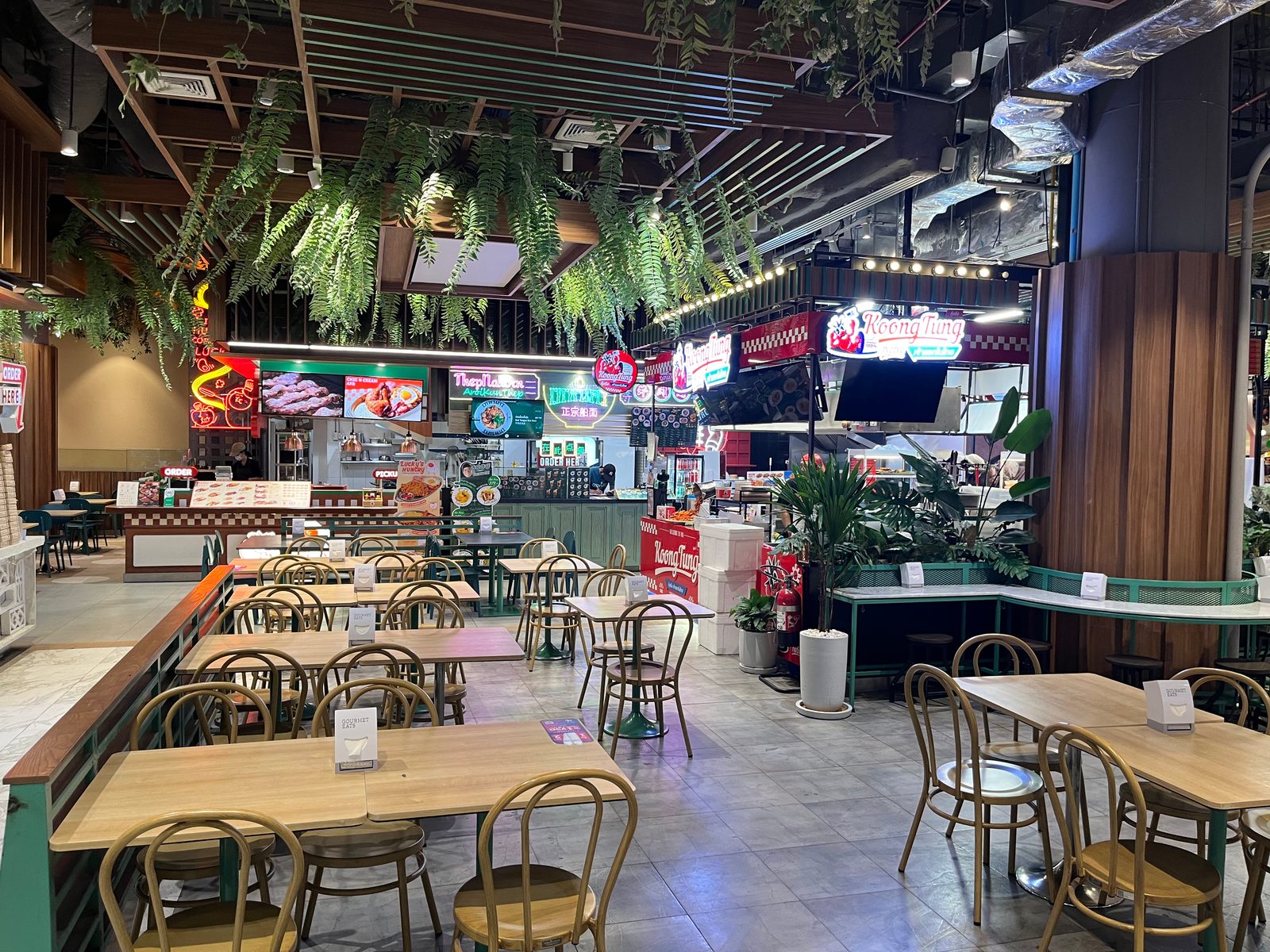 Empty tables under hanging plants in a Siam Paragon food hall, with neon stall signs behind