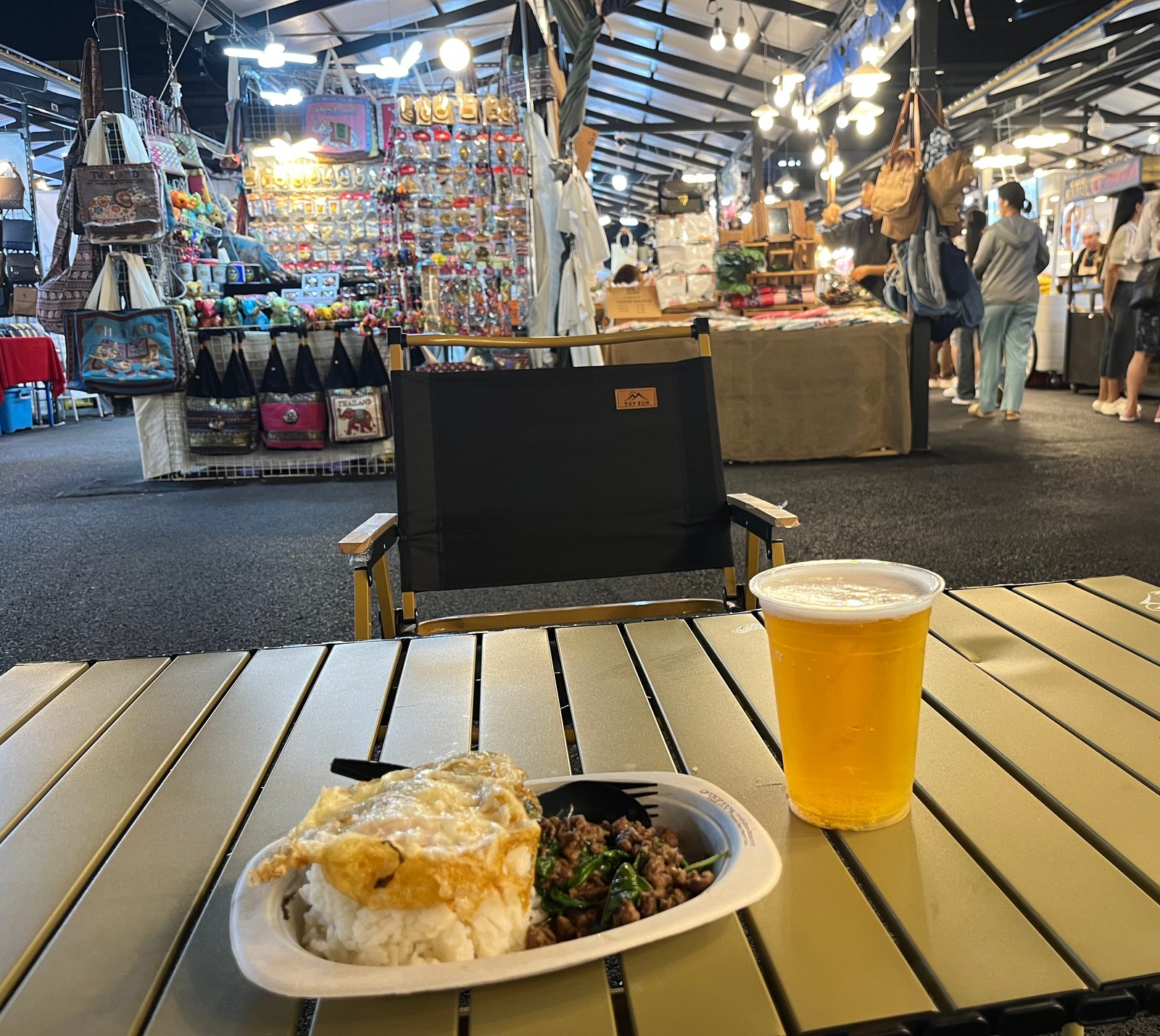 Rice with a fried egg and a basil stir-fry next to a beer, on a folding table at a night market