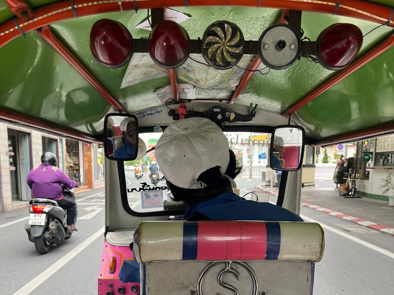 View from the back seat of a tuk-tuk on a Bangkok street, with the driver ahead and a scooter alongside