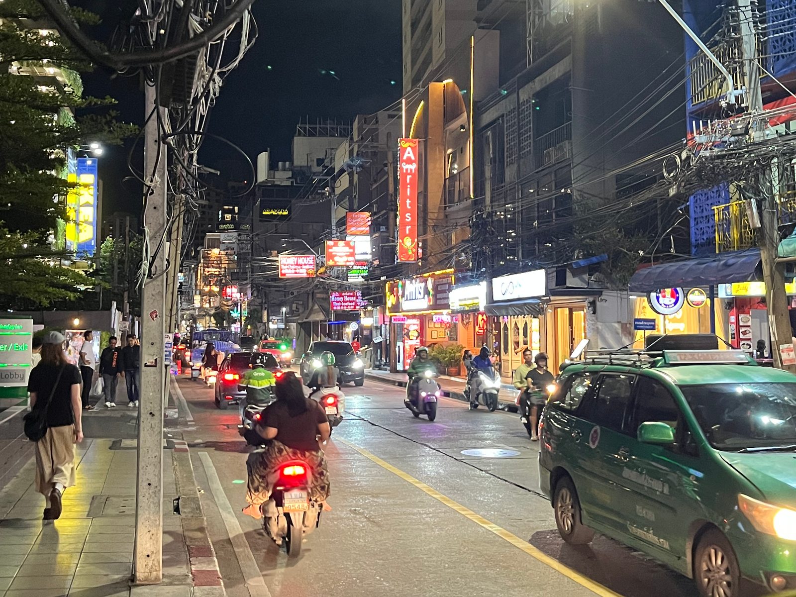 A lit street near Phrom Phong at night, with people on the sidewalk, motorbikes and a taxi