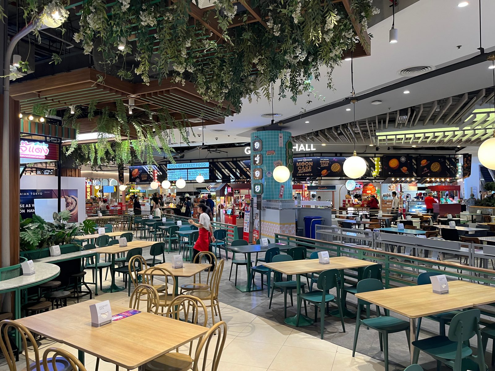 Rows of tables under hanging plants in a Siam Paragon food hall, with food stalls around